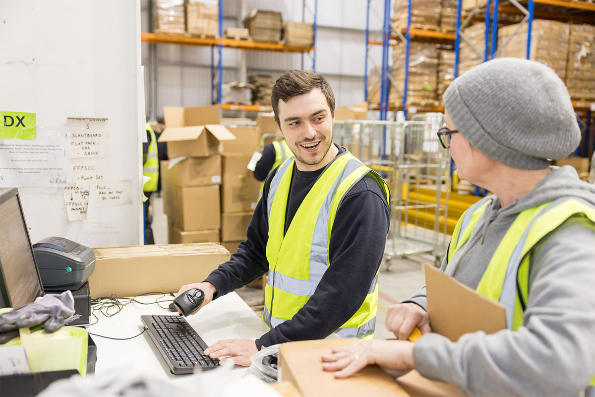 A man and woman wearing a high visibility vest talking to each other in a warehouse